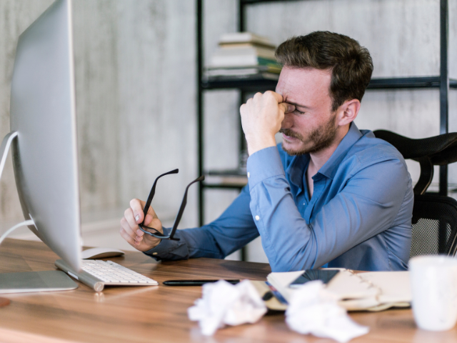 Homem sentado em frente ao computador, pressionando os olhos com os dedos e segurando os óculos, aparentando estresse ou problemas no trabalho.