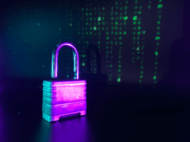 A imagem mostra um cadeado metálico iluminado por luzes em tons de roxo e azul, representando segurança e proteção. Ao fundo, aparecem caracteres verdes em padrão digital, reforçando o conceito de segurança cibernética e proteção de informações no ambiente virtual.