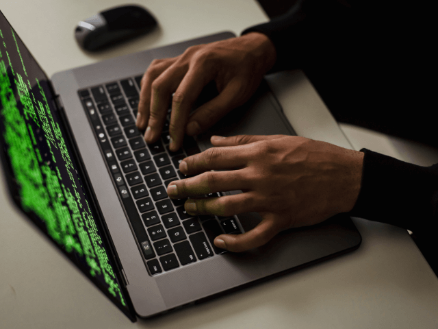 A imagem mostra uma pessoa digitando em um notebook, cuja tela exibe códigos em verde sobre fundo preto, lembrando interfaces de programação ou hacking. A cena transmite uma atmosfera de segurança da informação, programação ou atividades relacionadas à cibersegurança e análise de sistemas.