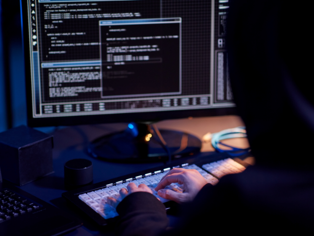 mostra uma pessoa de moletom escuro operando um computador com várias janelas de código e comandos na tela, transmitindo uma situação de programação ou possível invasão cibernética.