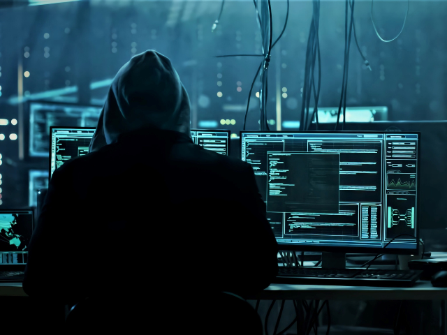 Pessoa com capuz sentada em frente a várias telas exibindo códigos, representando um hacker operando em ambiente escuro.