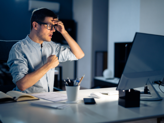 retrata um homem sentado à mesa, visivelmente preocupado ou surpreso ao olhar para a tela do computador, sugerindo que ele pode ter detectado um problema sério ou uma falha no sistema.