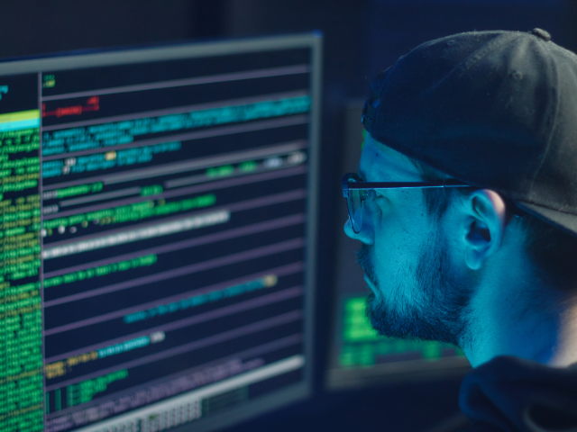 Um homem com boné e óculos observa atentamente múltiplos monitores com códigos de programação.