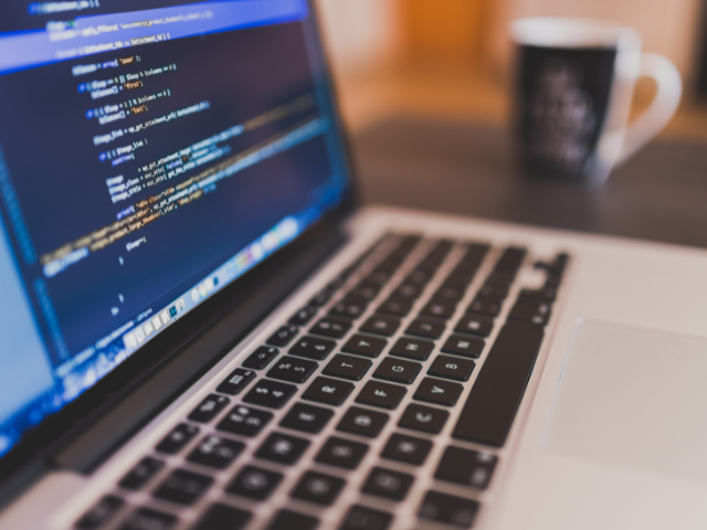  Tela de um notebook exibindo linhas de código com uma caneca de café desfocada ao fundo, remetendo ao ambiente de programação e desenvolvimento web.