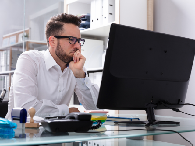 Homem de óculos e camisa branca observa atentamente o monitor com expressão séria, sugerindo preocupação com problemas técnicos ou falhas de segurança.